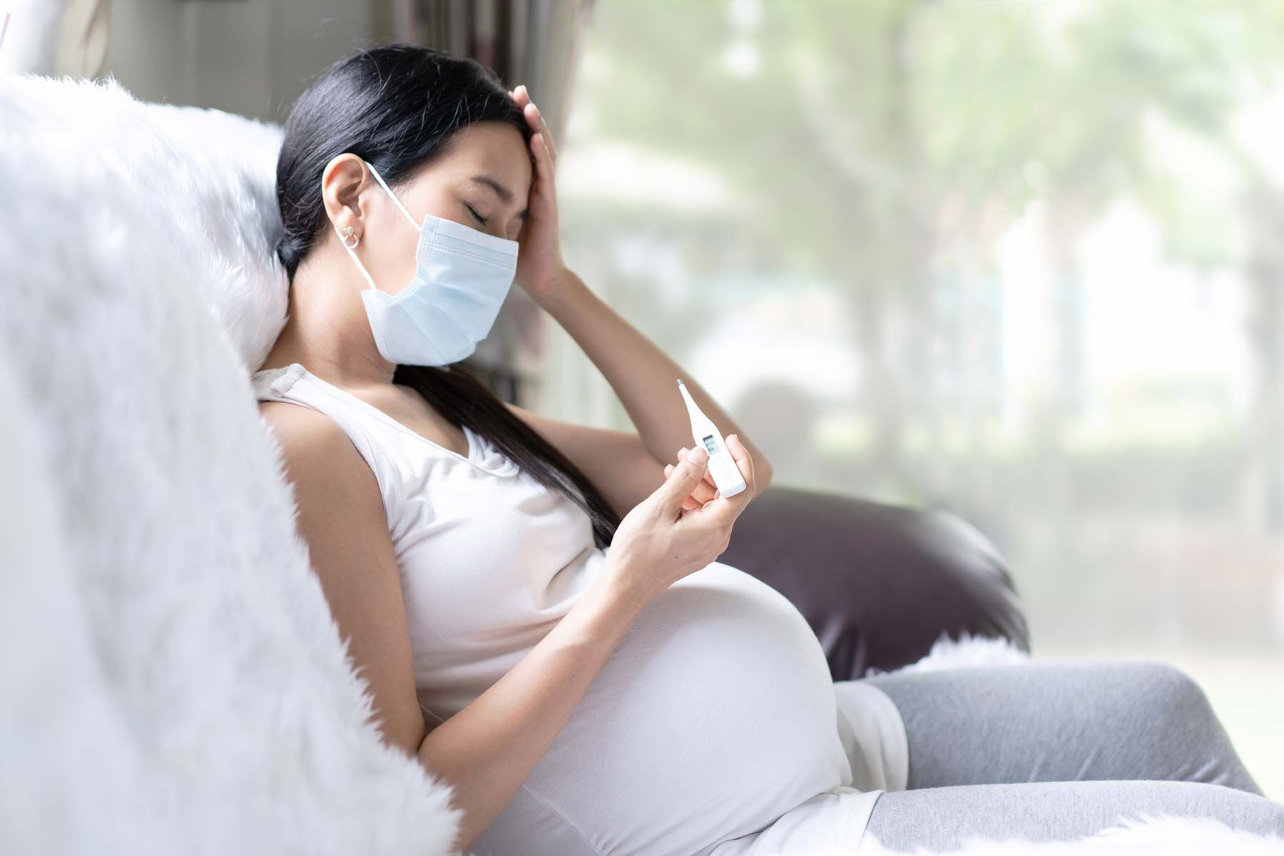 Pregnant woman with headache, taking temperature on sofa